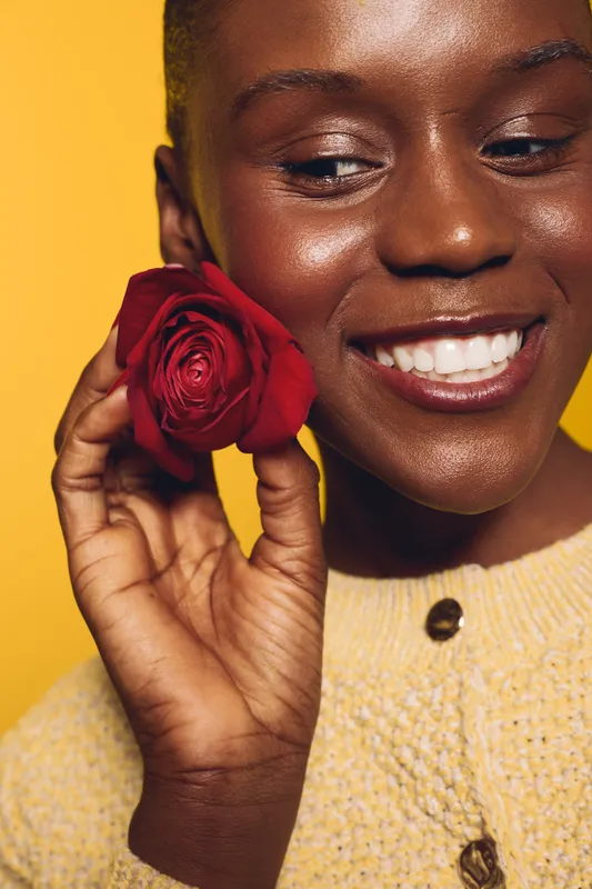 Close-up of a smiling black woman with dark skin holding a red rose near her face against a yellow background. Concept of beauty, happiness, and natural skincare with floral elements.