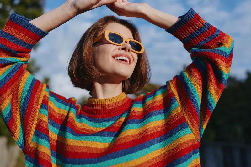 Woman in sunglasses and rainbow sweater smiling outdoors in a vibrant portrait, short hair and raised arms enjoying sunlight and carefree mood in colorful casual fashion.
