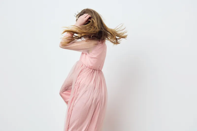 Young woman in a flowing pink dress, joyfully twirling with her hair in motion against a minimalist white background, embodying freedom and elegance