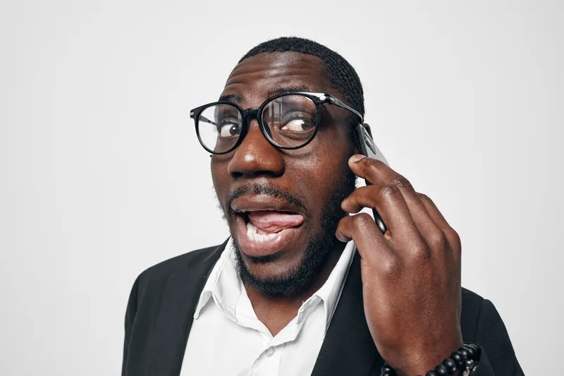 Black man with dark skin and glasses talking on smartphone with expressive face in white shirt and black jacket on white background. Communication, technology and emotion.