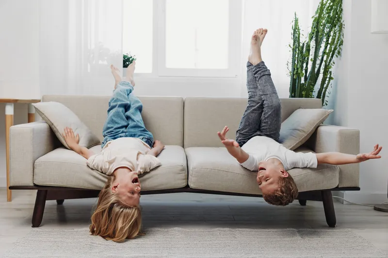 Two children laying upside down on a beige sofa in a bright living room with large windows and plants. They laugh and play, expressing joy and carefree childhood moments indoors.