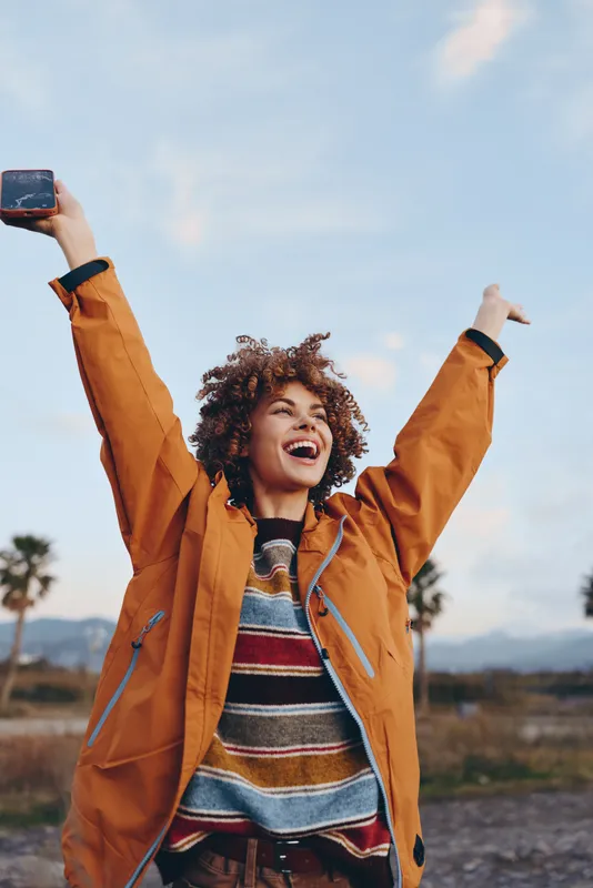Smiling woman lifestyle outdoors in orange jacket holds a smartphone above her head for a selfie, arms raised in joy, curly hair in wind, open sky landscape, positive mood today