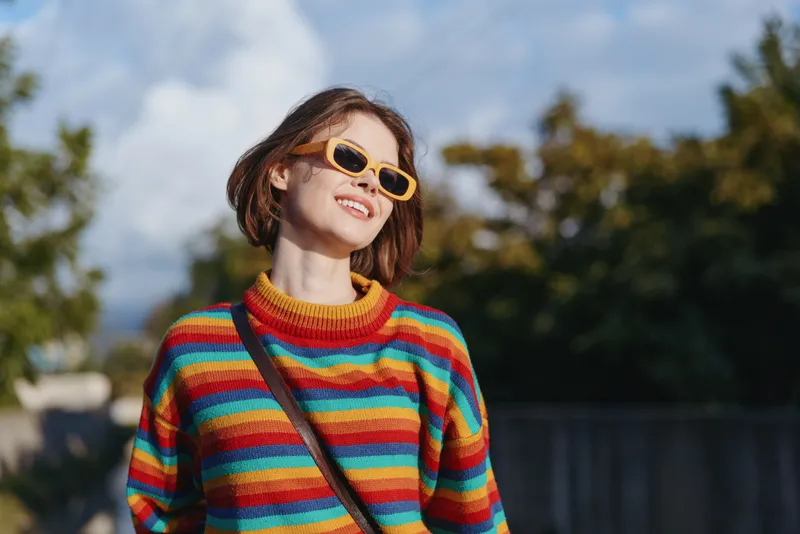 Young woman in her early 20s wearing a colorful striped sweater and yellow sunglasses, smiling in an outdoor urban park with a crossbody bag, casual outfit for travel lifestyle and sunny mood