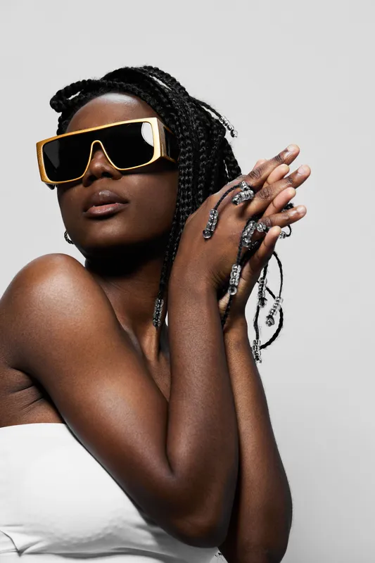 Stylish African model with braids, wearing oversized sunglasses and elegant jewelry, posing with a confident and bold expression against a minimalist gray background