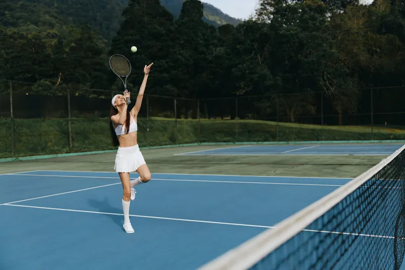 Young female tennis player in a white outfit serving the ball on a sunny day, showcasing determination and focus on a vibrant blue court surrounded by greenery