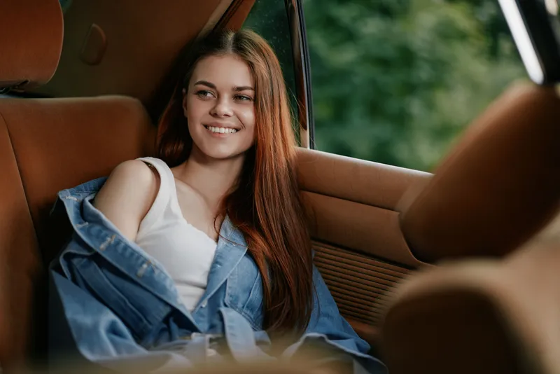 Young woman with long hair in a denim jacket smiling in a cozy car interior, capturing a carefree moment in nature, evoking feelings of joy and freedom.