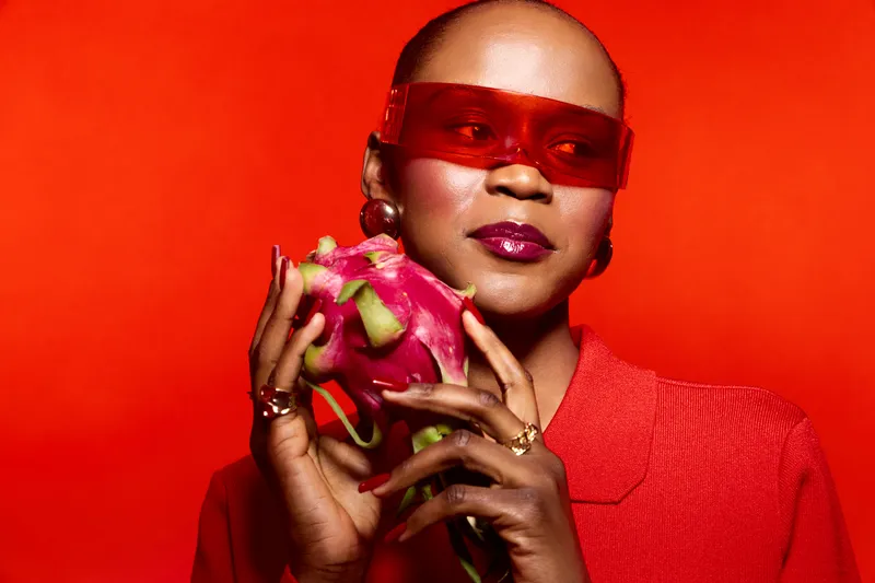 Portrait of confident black woman with dark skin wearing red sunglasses and clothing holding a dragon fruit. Vibrant red background highlights fresh fruit and stylish accessories concept.