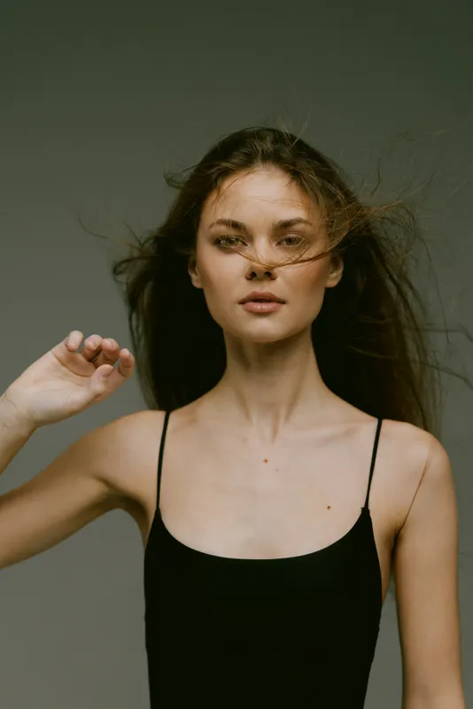 Serious woman in black sleeveless dress with windblown hair posing against neutral background