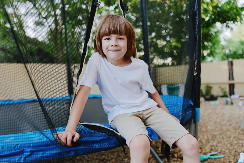 Happy boy sitting on a trampoline, enjoying summer outdoors, smiling brightly amidst a playful background of green trees and soft sunlight.