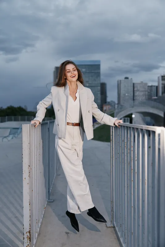 Confident woman in beige business suit walking outdoors on urban bridge with gray sky behind, smiling in modern fashion style