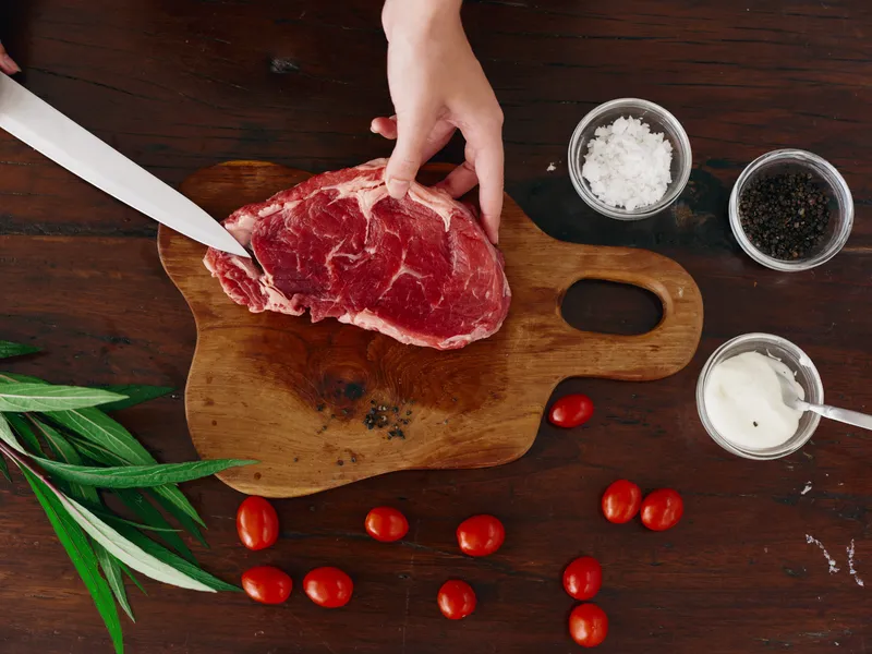 Женщина с ножом в руке нарезает свежее стейковое мясо для жарки на кухне