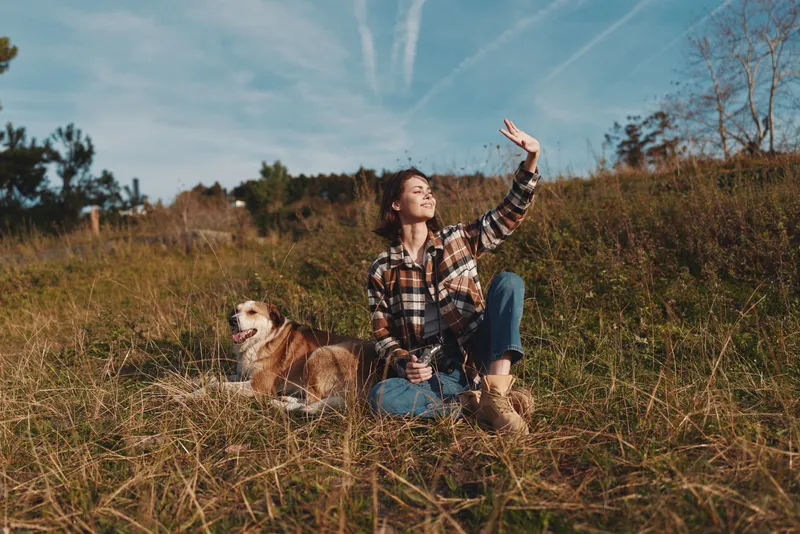 Woman in plaid shirt sits in a grassy field with a dog beside her, raising her arm toward the bright sky during a sunny outdoor moment in nature today.
