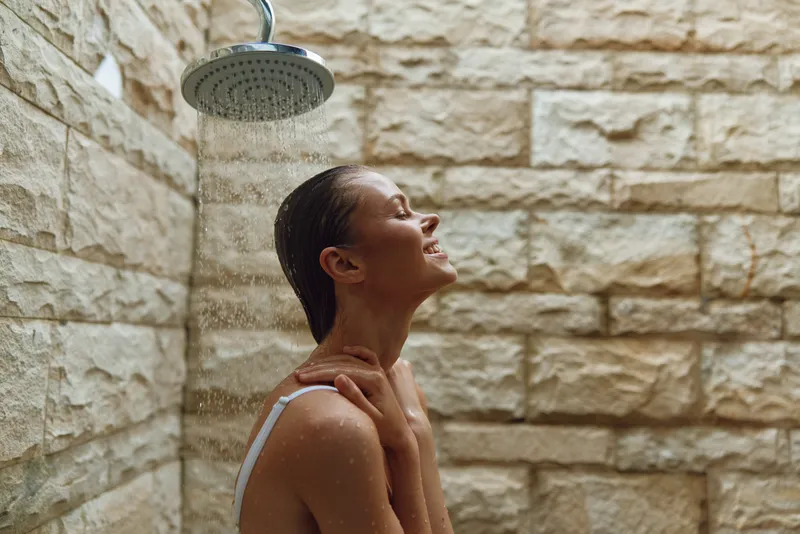 Relaxed woman enjoying outdoor shower, wet hair, stone wall background, natural light, refreshing moment, peaceful expression, summer wellness.