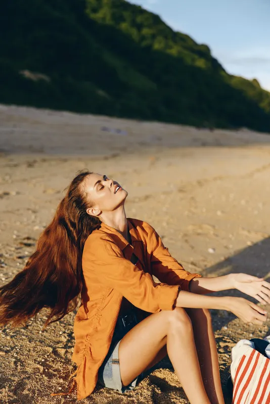 Young woman with long brown hair enjoys the warm sunlight while sitting on a sandy beach wearing an orange shirt and denim shorts, feeling relaxed and peaceful.