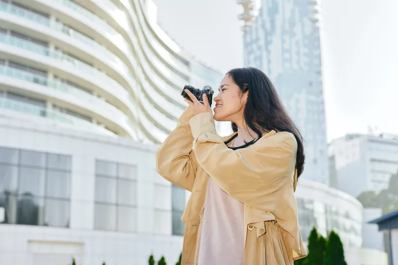Woman with camera in an outdoor urban setting, capturing a candid moment near modern architecture, daylight, city life, travel vibe, photography focus and cityscape backdrop