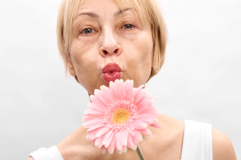 Woman closeup pink gerbera flower portrait beauty studio white background soft light lips pursed holding flower near face playful calm spring mood gentle skin tone