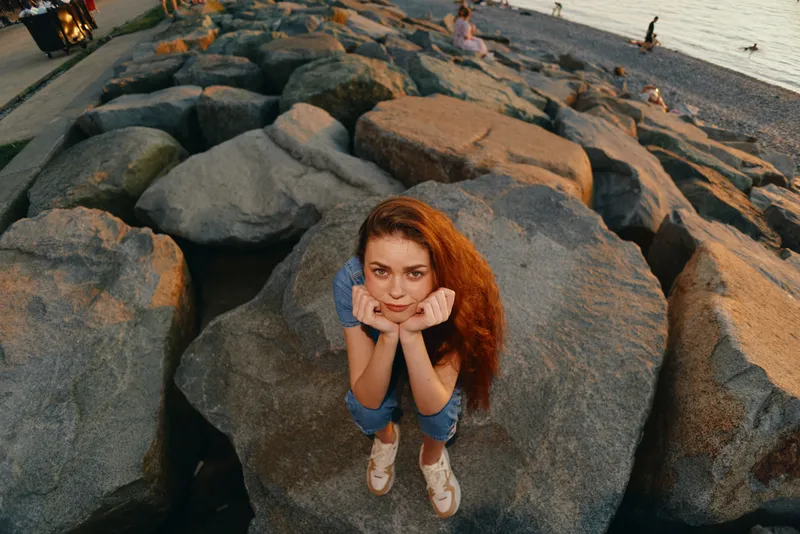 lifestyle woman with red hair in modern boho-western denim street style standing on rocks by the beach in warm film color lighting, casual outfit, relaxed mood
