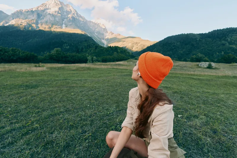 Woman in outdoor landscape sits in a green field beneath mountains, wearing a bright orange beanie, enjoying nature and a calm mood in a casual lifestyle image