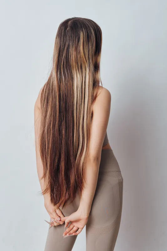 Woman with long straight hair standing with hands behind back wearing casual beige top and leggings in front of plain light background for beauty and style concept.