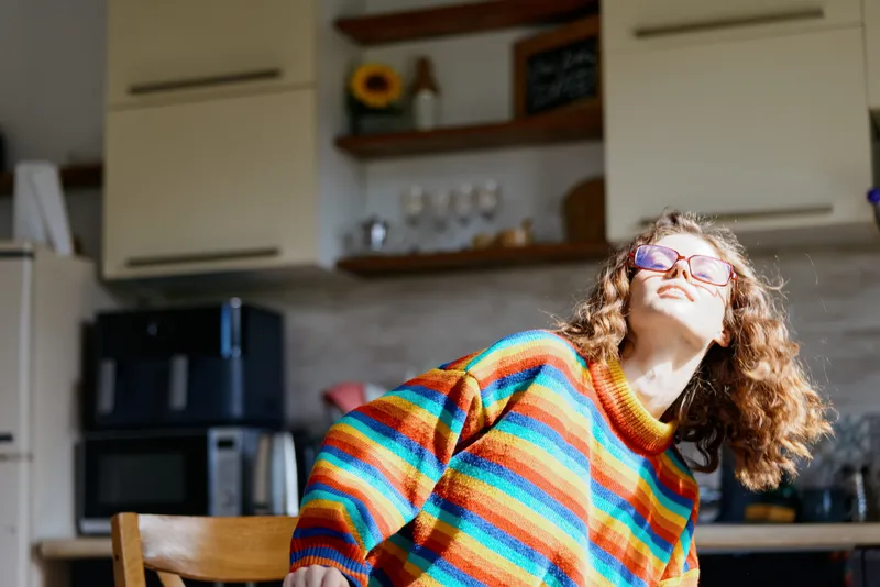 Bright kitchen scene features a smiling woman in a colorful striped sweater, looking upward with joy, capturing a casual home lifestyle and a candid warm indoor moment.