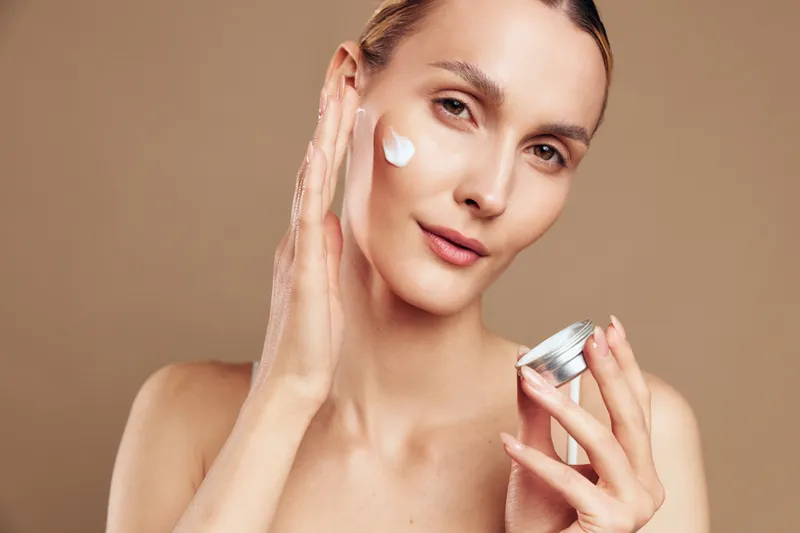 Fresh faced woman applies moisturizer to her cheek with a dab of cream, close up portrait, smooth skin, soft lighting, calm skincare routine for healthy glow and self care