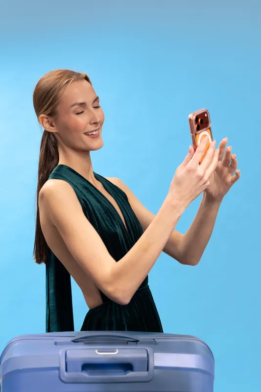 Young woman in elegant dark dress takes selfie with mobile phone standing behind blue suitcase on blue background. Concept of travel, vacation, and modern lifestyle.