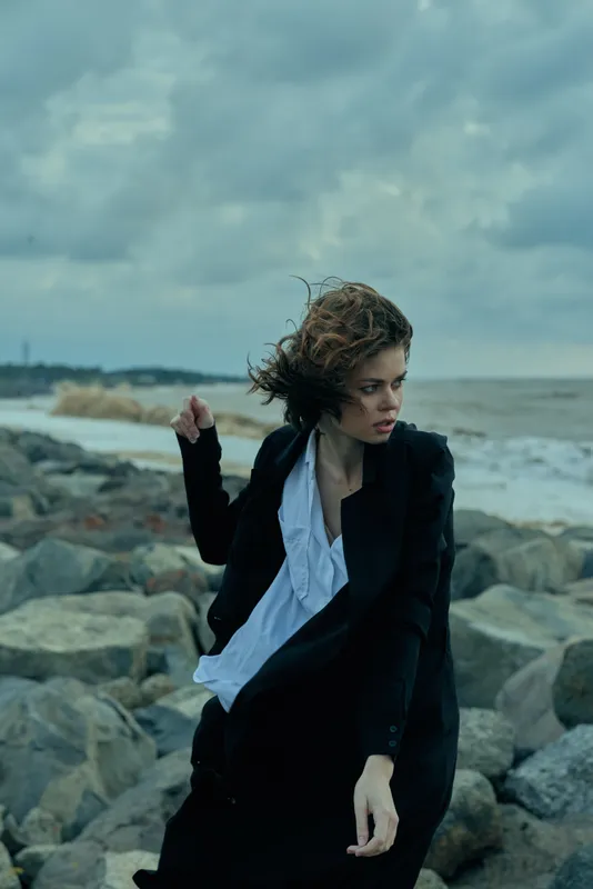 Fashion conscious woman stands on a windy rocky shore, blazer and loose shirt billowing, capturing moody coastal drama and bold style.