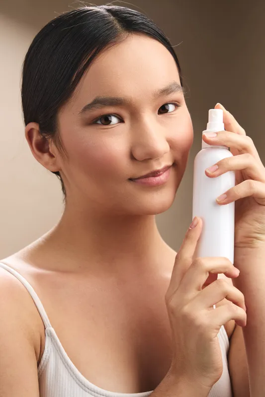 Asian woman smiles softly as she holds a spray bottle in a neutral setting, presenting a calm skincare moment and a modern personal care routine with a subtle, fresh ambience.