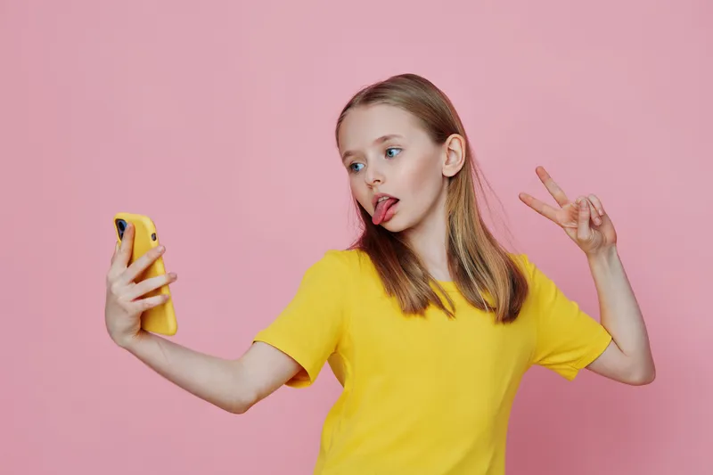 Playful girl taking a selfie with a smartphone, showcasing fun emotions in a vibrant yellow t shirt against a pink background