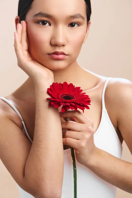 asian woman gazes softly at the camera as she holds a vibrant red flower near her chest, studio lighting and a clean backdrop create an elegant, serene fashion portrait.