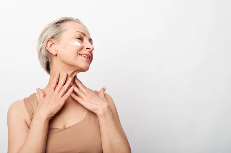 Smiling mature woman with short blonde hair applies cream on face and neck, wearing beige sleeveless top. Concept of skincare, aging gracefully, self-care, and natural beauty on white background.