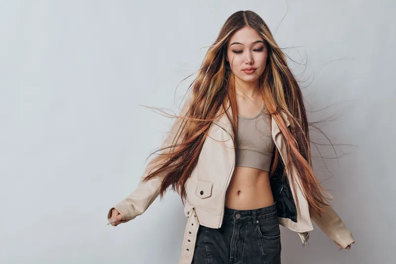 Woman with long hair wearing beige jacket and crop top posing against plain white wall with eyes closed and loose hair for fashion and beauty concept.