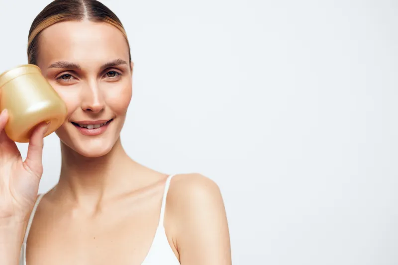 Smiling woman in closeup portrait applying face cream and serum from a gold bottle, showcasing skincare, beauty, wellness and personal care on a clean white background.