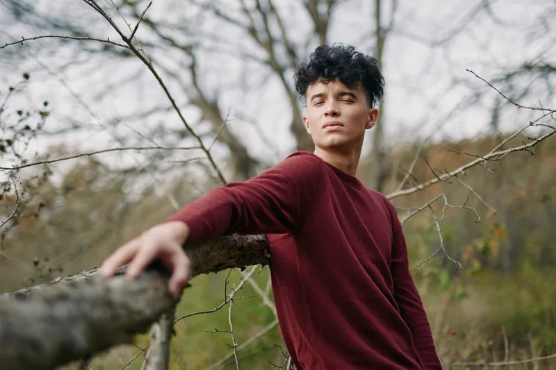 A candid portrait of a young man in a maroon sweater leaning on a wooden fence, radiating authenticity and quiet credibility in a calm natural setting.