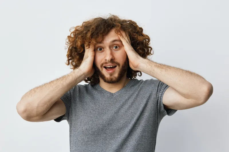 Surprised male model with curly hair expressing excitement against a neutral background, showcasing joy and spontaneity in everyday life