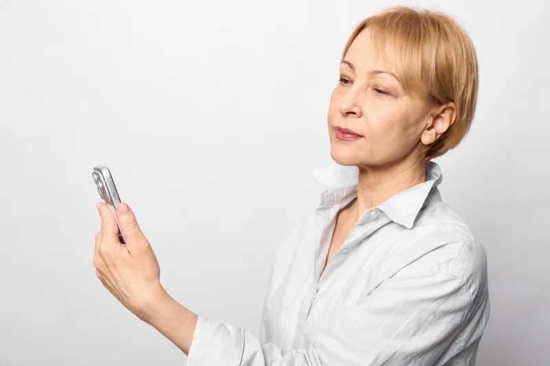 Elderly woman with short hair holding a smartphone, studio portrait on a white background. Communication, technology, and social connection in an indoors calm moment.