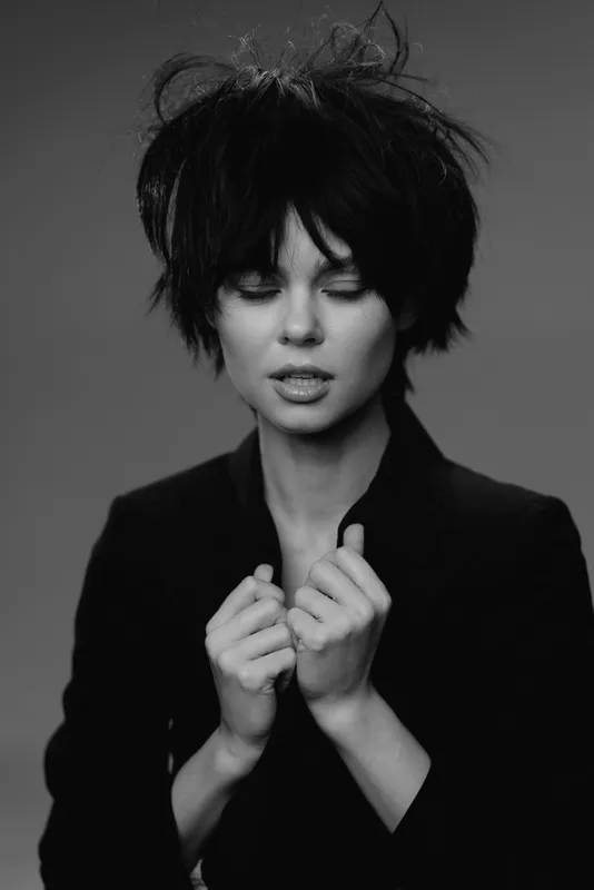 Black and white portrait of thoughtful woman with short messy hair in dark clothing expressing calm emotion.
