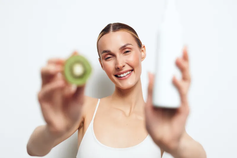Smiling woman demonstrates skincare routine with kiwi and cream, shown in a bright studio setting. Clear focus on facial care, natural beauty, and healthy skin.