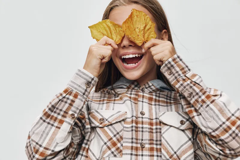 Young woman with long blonde hair smiling and holding two autumn leaves in front of her eyes wearing a plaid shirt on a light background.