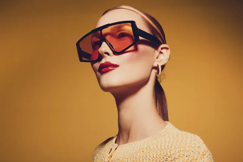 Stylish woman with slicked back hair wears oversized orange sunglasses and red lipstick against a warm yellow background. Concept of fashion, beauty, and modern accessories.