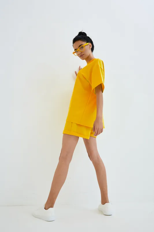 woman in yellow outfit posing confidently against white background, wearing casual summer clothing and stylish glasses, minimalistic fashion concept