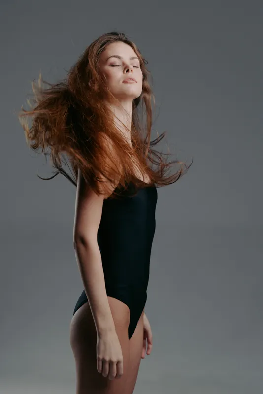 Young woman in black bodysuit with flowing hair posing confidently against gray background.