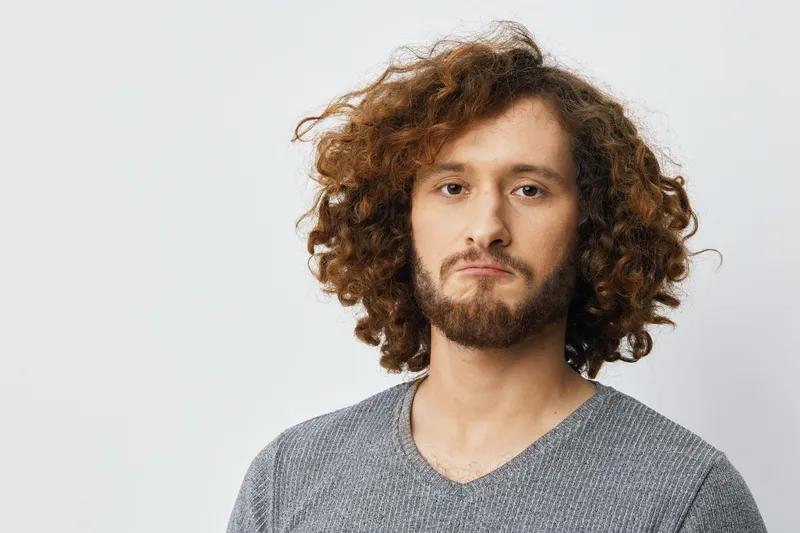 Young man with curly hair and beard, expressing a thoughtful emotion against a light background, perfect for concepts related to self reflection, identity, and emotions