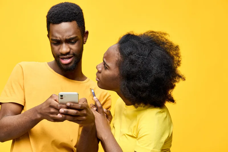 A young Black couple displaying surprise and curiosity while looking at a smartphone against a vibrant yellow background, showcasing modern communication and emotions
