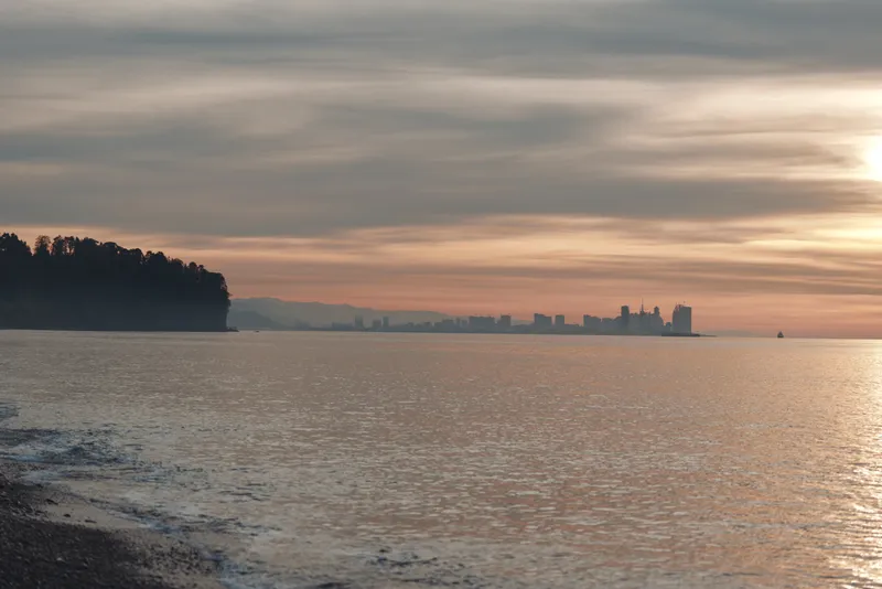 Sunset over calm water with distant city skyline and silhouettes along the shore, warm orange sky reflecting on gentle waves, a tranquil scene for travel, nature, and evening mood.