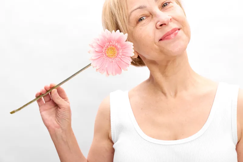 Smiling woman with blonde hair holds a pink flower on a wooden stick in a studio, white background, spring mood, wellness portrait, soft light