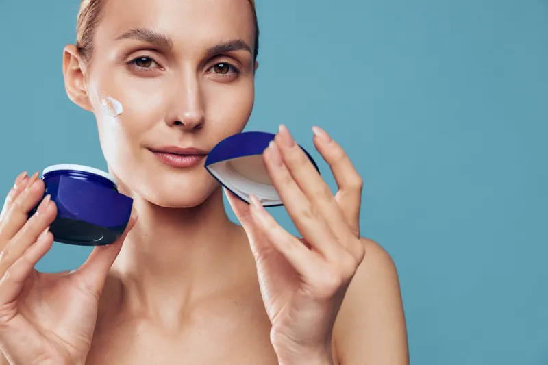 Caring woman applies facial cream on her cheek with a blue background, showcasing skincare routine, hydration, natural glow, closeup, calm cosmetics moment, selfcare vibe