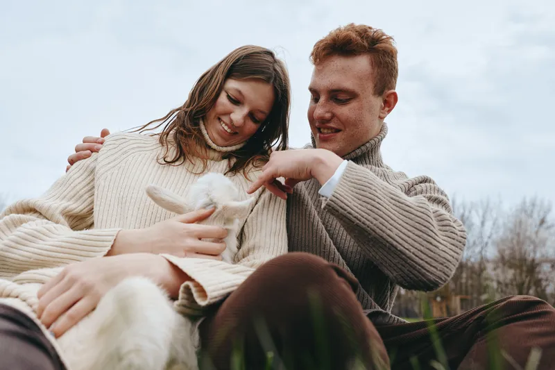 Lifestyle moment with a joyful couple outdoors, wearing cozy sweaters, a gentle goat kid resting on lap, warm natural tones, soft breeze, candid affection, relaxed setting, greens and blue sky