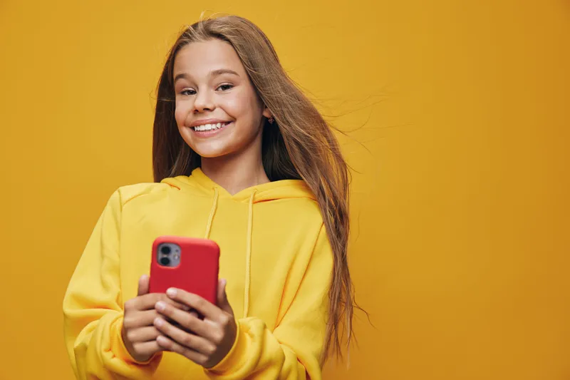 Teenage girl smiling and holding smartphone in red case wearing yellow hoodie against yellow background, casual technology use and happy expression with long hair.
