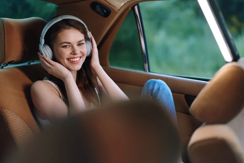 Young woman enjoying music in a car with headphones, smiling joyfully in a relaxed setting amidst lush greenery.